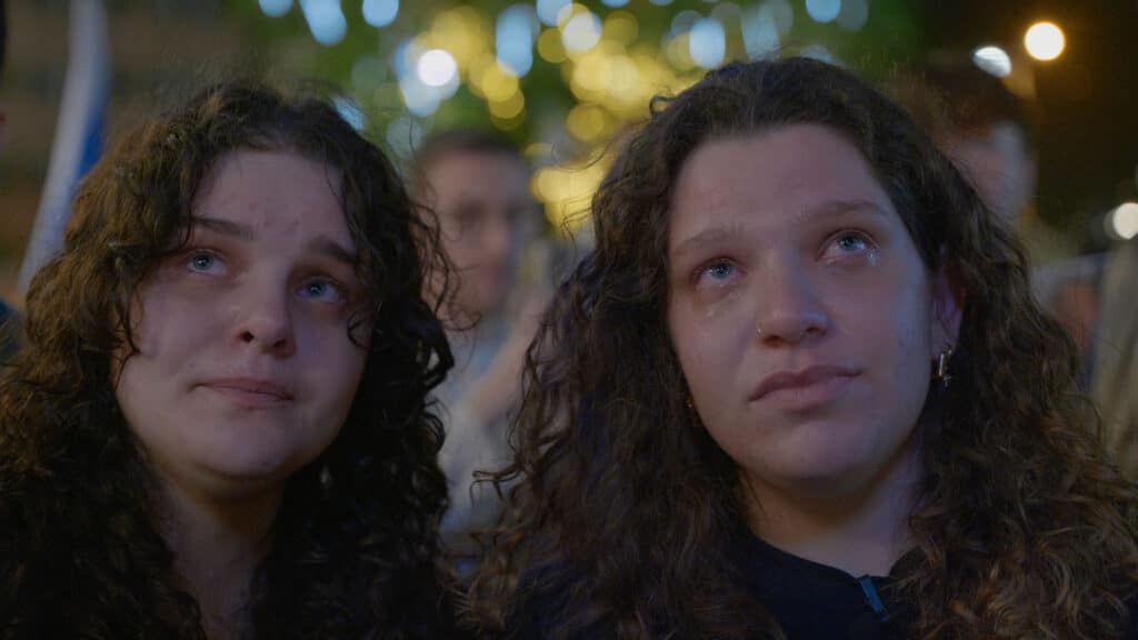 Two women with curly hair stand side by side at night, both with teary eyes and somber expressions. Blurred lights and people are visible in the background, creating an emotional and reflective atmosphere.