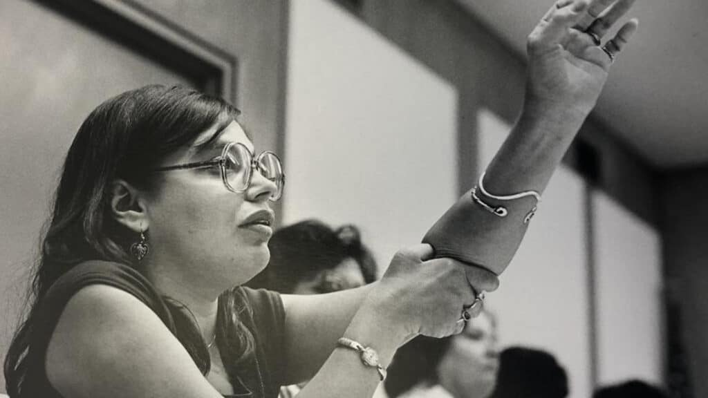A woman with glasses raises her arm and holds her elbow while speaking or asking a question in a classroom or meeting setting. Other people are blurred in the background.